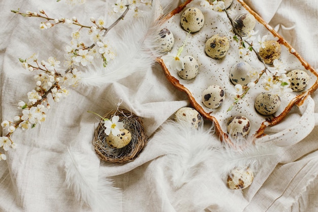 Rustic easter flat lay Stylish easter eggs and blooming spring flowers on linen fabric Happy Easter Natural quail eggs in tray feathers and cherry blossoms on rural table