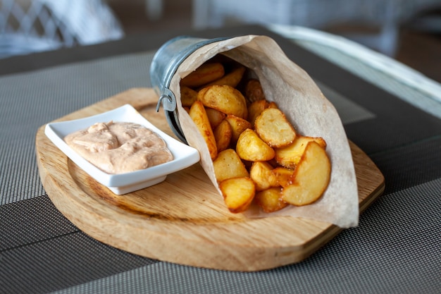카페의 야외 테라스에 있는 나무 접시에 소스와 함께 종이 봉투에 소박한 튀긴 감자 웨지.