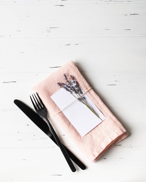 Rustic romantic table setting with paper card, pink napkin, lavender and appliances on white wood table. Top view. Copy space