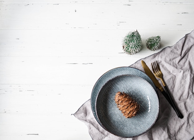 Rustic table setting for christmas or new year. Blue plates, grey napkin, pine cone and appliances on white wood table. Top view. Copy space