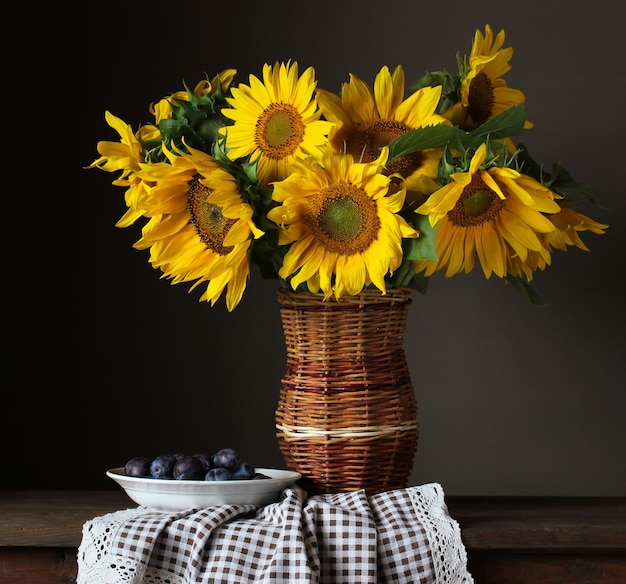 Rustiek stilleven zonnebloemen in een rieten vaas en een bord met pruimen