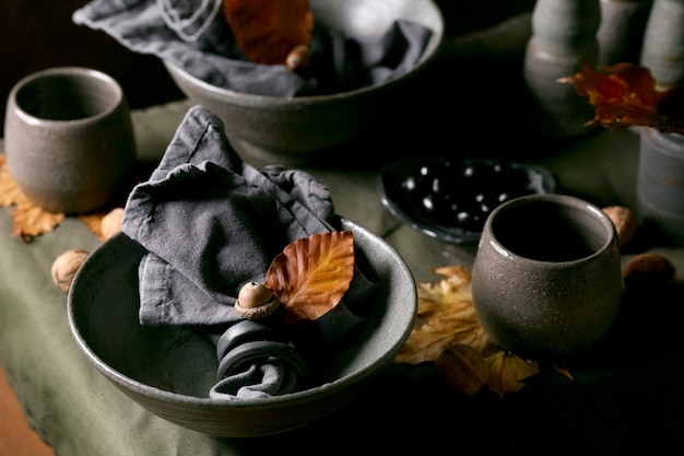 Rustieke herfst Halloween- of Thanksgiving-tafel met leeg ambachtelijk keramisch servies, grijze ruwe kommen en kopjes op linnen tafelkleed met herfstgele bladeren en eikels. Donkere achtergrond