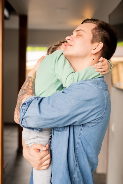 Foto rustige liefdevolle hipster jonge vader met tatoeage op arm ogen gesloten houden terwijl schattige dochter knuffelen en haar troosten