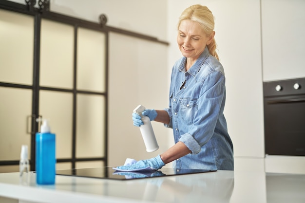 Rustige rijpe blonde vrouw in rubberen handschoenen die oppervlakken in de keuken schoonmaakt met een doek en spray