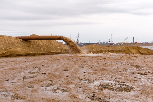 Ржавая труба на берегу реки, из которой стекают сточные воды, грязная песчаная жижа