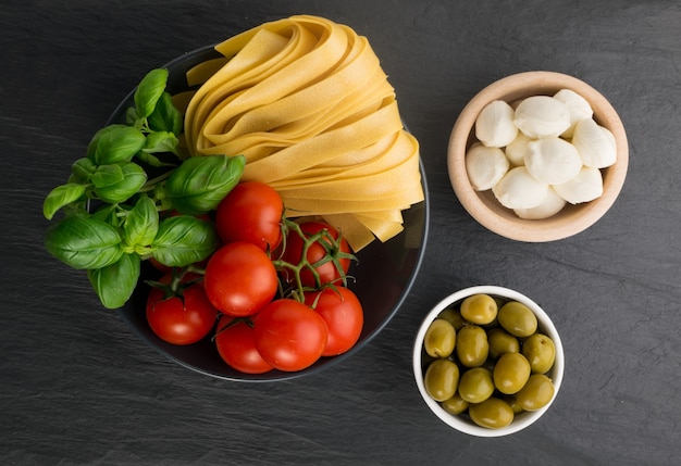 Ruwe gele Italiaanse pasta pappardelle, fettuccine of tagliatelle bovenaanzicht. Ei zelfgemaakte noedels, langgerolde macaroni of ongekookte spaghetti met olijven, tomaten, basilicum en mozzarella kaas