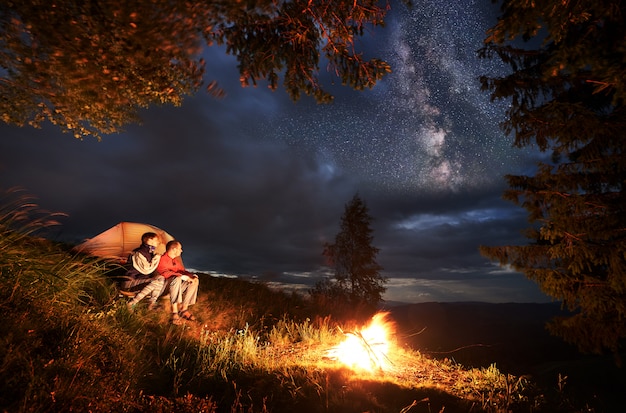 's nachts kamperen. jong koppel wandelaars zitten bij het kampvuur en verlichte oranje tent, op zoek in de verte onder de avondlucht. door wolken aan de hemel zijn heldere sterren zichtbaar.