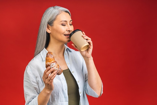 'S ochtends koffie om te gaan Charmante senior vrouw permanent met papieren kopje warme drank en croissant geïsoleerd op rode achtergrond voorbereiden op goede werkdag Rijpe zakelijke dame neemt een koffiepauze