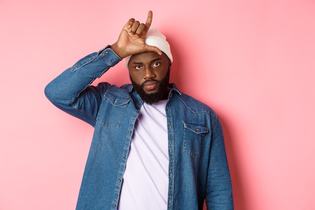 Sad african-american man showing loser sign on forehead and staring at camera, standing over pink background