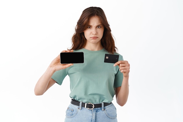 Фото Грустная и мрачная девушка, хмурясь, расстроена, показывая горизонтальный пустой мобильный экран и пластиковую кредитную карту, жалуясь стоя на белом фоне