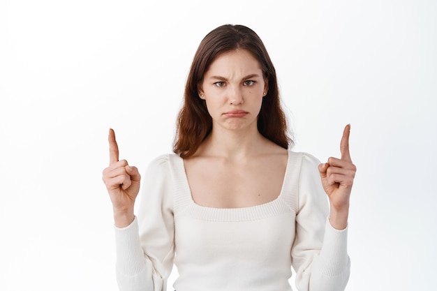 Sad and angry young woman pointing fingers up, blame you, sulking and frowning displeased, showing something unfair or bad, complaining, standing against white wall