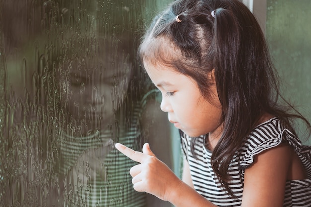 Sad asian little girl looking outside through the window in the rainy day