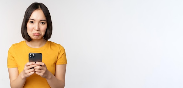 Sad asian woman holding smartphone looking upset with regret standing in yellow tshirt against white background