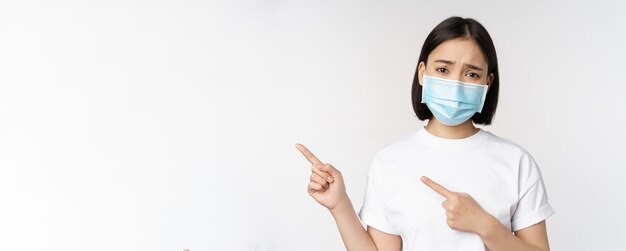 指を指している医療マスクの悲しいアジアの女性は、白い背景の上に立っているバナーを示す眉をひそめ、動揺して不平を言っている