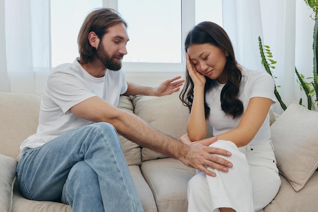 a sad asian woman talks to a man in tears at home sitting on the couch young couple of different nationalities and conflict of interest in a couple