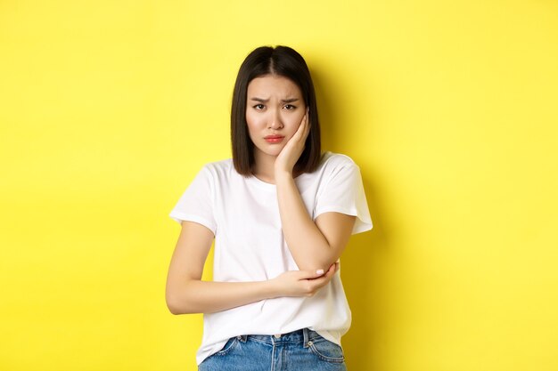 頬に触れて眉をひそめ、歯痛を抱えている悲しいアジアの女性は、黄色の背景に対して暗い立っている歯科医を見る必要があります。