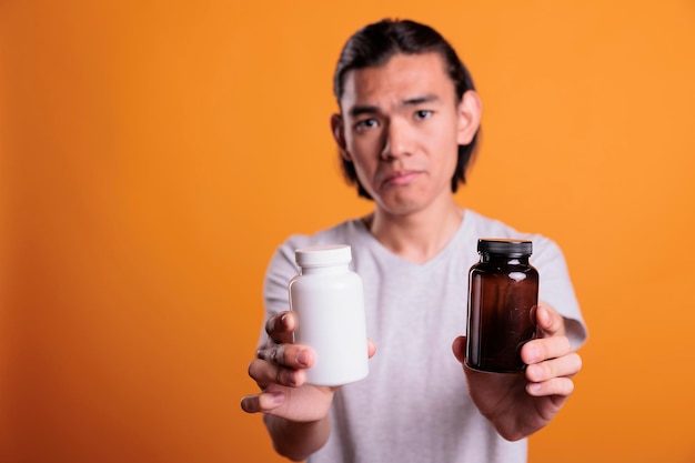 Sad asian young man taking pills, drugs bottles close up. Disease treatment, illness remedy, upset patient holding medicaments, person showing vitamins, supplement capsules