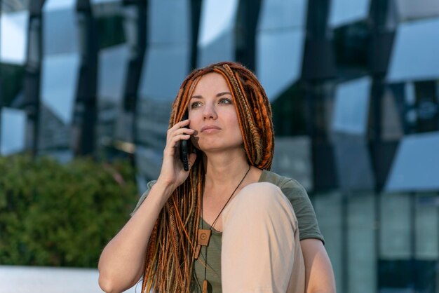 Sad beautiful red-haired girl with dreadlocks is talking on the phone. Portrait on the background of an office building