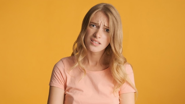 Sad blond girl with frowning face posing on camera over yellow background