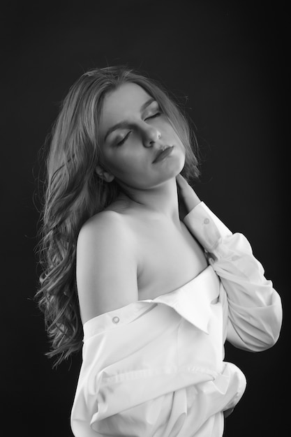 Sad blond woman wears men's shirt posing in the shadows at studio. Black and white image