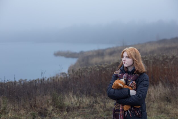 Sad blonde teenager woman hugging teddy bear by foggy lake. Concept of adolescence and adolescent problems.
