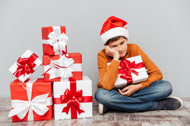 Sad bored little boy in santa claus hat sitting near present boxes