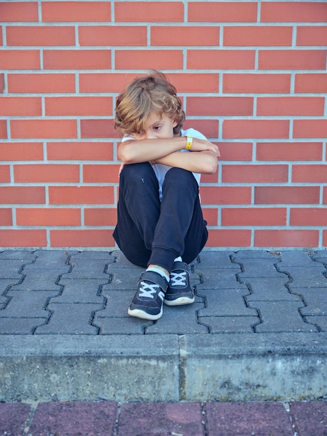 Sad boy hugging knees and sitting at brick wall on street