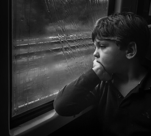 Photo sad boy looking through window during rainy season