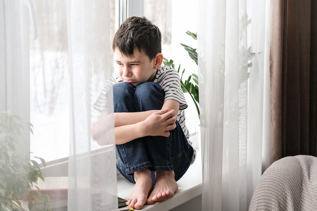 悲しい少年が膝を抱いて窓辺に座っている機嫌が悪いうつ病少年は家で一人で悲しい