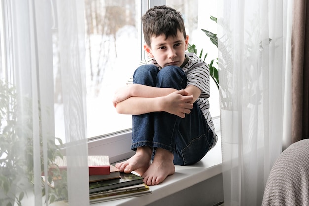 A sad boy sits on the windowsill hugging his knees Bad mood depression The boy is sad alone at home