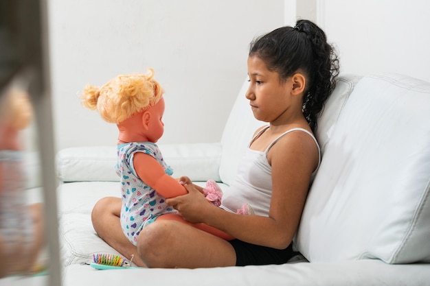 Sad brown girl sitting next to her doll on the living room sofa\
looking at the doll