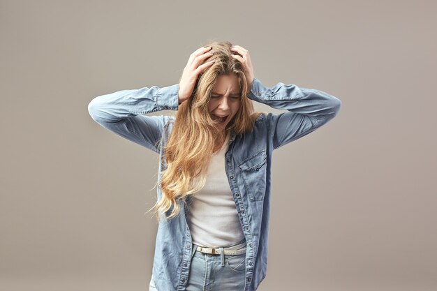 Sad brown-haired girl dressed in a white t-shirt and jean holds her hands on her head and shouts on a gray background .