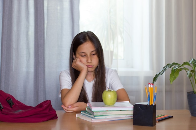 Sad brunette girl sits at home over books on which there is an apple back to school concept