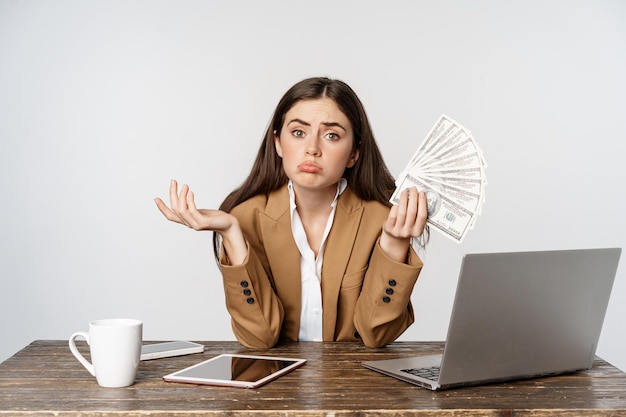 Sad businesswoman sitting in office with laptop holding money and looking disappointed in income bus...