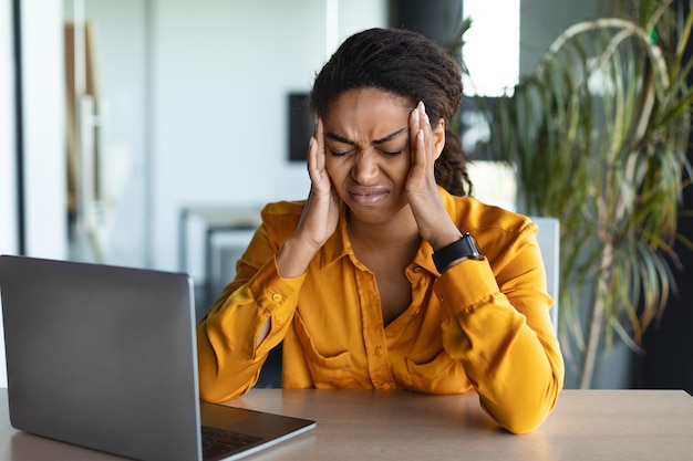 Sad businesswoman sitting with laptop at workplace suffering from headache touching temples working in office