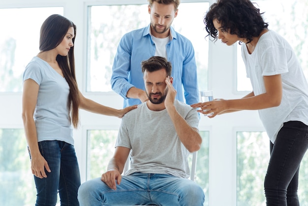 Sad cheerless bearded man holding his temple and suffering from the headache while being surrounded by friends