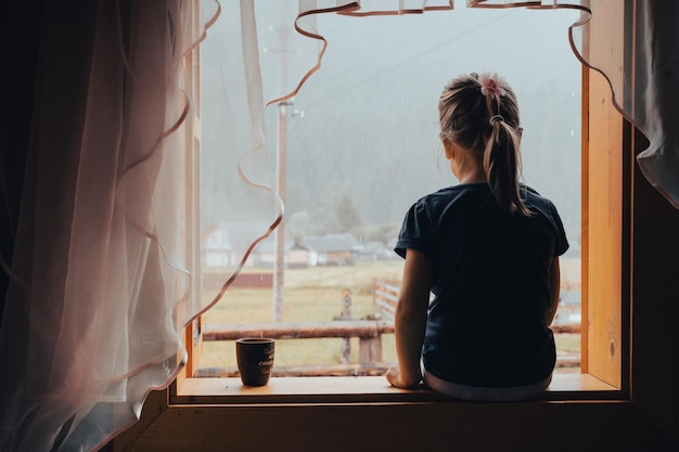 Sad child looking through rainy window at home