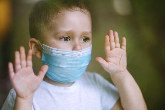 Sad child in a medical mask looks out the window Stay at home