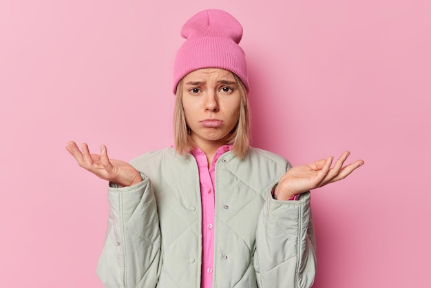 Sad clueless European woman shrugs shoulders with hesitation faces difficult decision feels unsure wears hat and jacket isolated over pink background. People hesitation and confusion concept