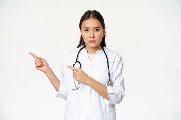 슬프고 걱정스러운 여성 의사는 왼쪽 손가락을 가리키며 중요한 프로모션 정보를 보여주며 흰색 배경 위에 서 있습니다.