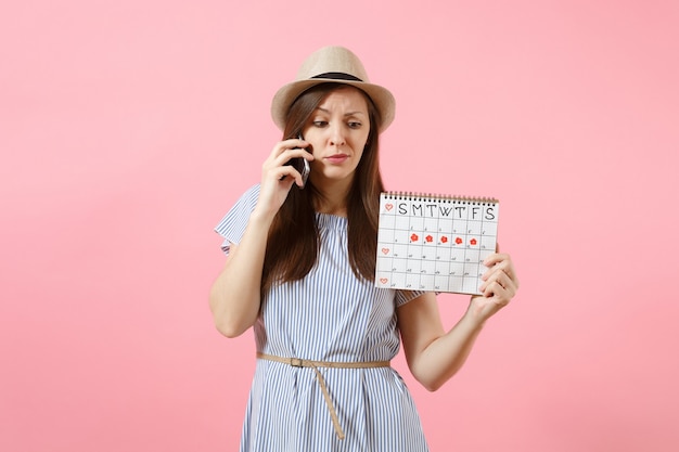 Sad confused woman talking on mobile phone, holding periods calendar for checking menstruation days isolated on bright trending pink background. Medical, healthcare, gynecological concept. Copy space.