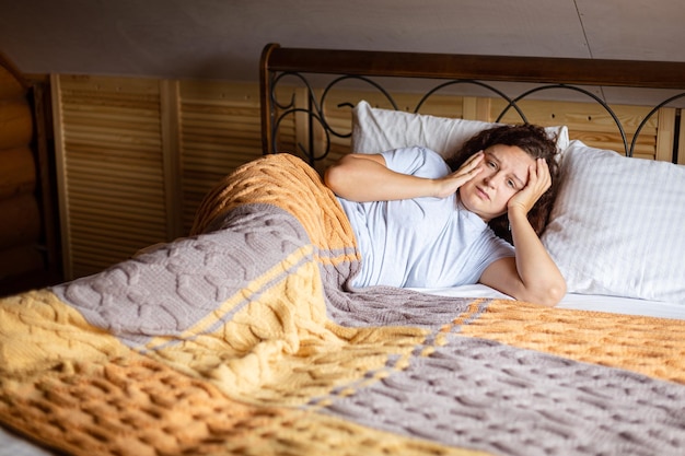 Triste donna bruna riccia sdraiata sul letto sotto le coperte di spugna con la faccia preoccupata che ha problemi interiori indossando il pigiama a casa durante il giorno momento di ansia nella vita adulta