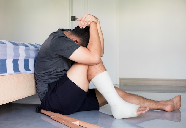 Печальный депрессивный азиатский мужчина страдает возле кровати он сидит и касается его лба и стресс с симптомами сломанной ноги и белого гипса неожиданные несчастные случаи страхования от несчастных случаев
