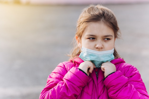 Sad, depressed little girl in sterile medical protective from virus mask outdoors. Health care, epidemic, pandemic, illness concept