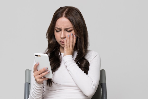 Sad depressed young woman reading phone message phone, sitting on chair, isolated. Unpleasant news, love depression