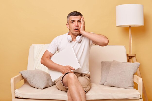 Sad despair shocked adult man wearing white tshirt and shorts sitting on sofa with closed laptop against beige wall showing facepalm having job problems