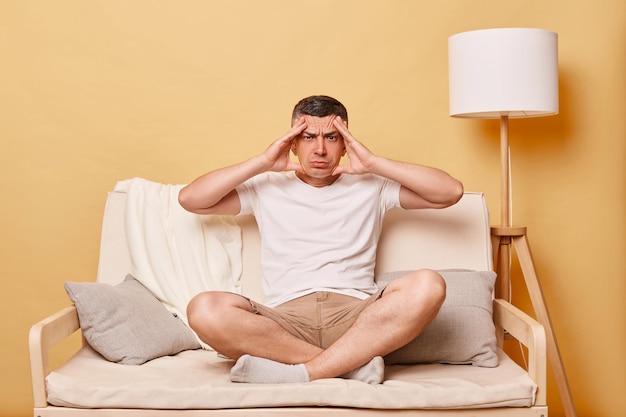 Photo sad despair sick ill brunette young man wearing casual white t shirt sitting on sofa against beige wall suffering terrible headache massaging temples