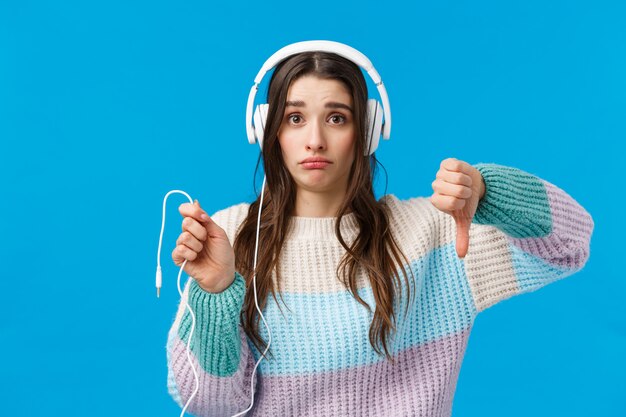 Sad and displeased, gloomy cute brunette woman in big headphones, grimacing disappointed, showing thumbs-down and holding dropped wire of earphones, standing blue 