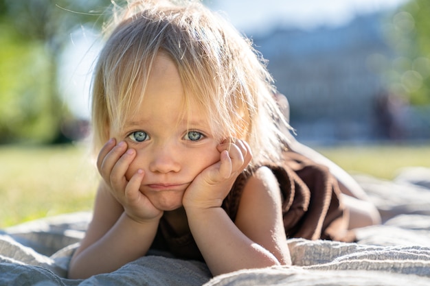 Sad displeased little child girl with blue eyes looking at camera, lying on plaid on grass, outdoor.