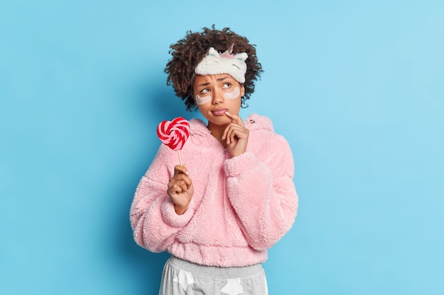 Sad displeased woman looks pensively aside holds delicious lollipop going to sleep applies hydrogel patches under eyes isolated over blue wall